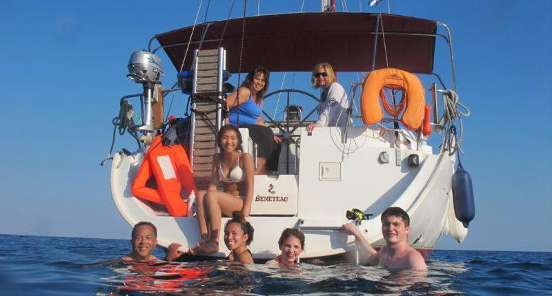 A group of friends enjoying a skippered yacht charter, with some swimming in the sea and others relaxing on the back of a white sailboat under a clear blue sky. The perfect sailing experience for a summer adventure