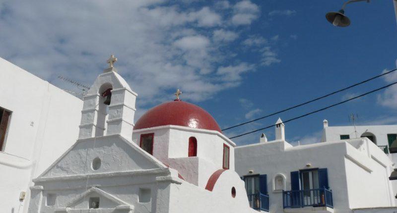 A traditional white-washed church with a red dome and bell tower, set against a bright blue sky with scattered clouds. The church is surrounded by white buildings with blue doors and windows, characteristic of Greek island architecture.