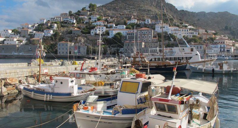 A scenic view of Hydra's harbor, filled with traditional fishing boats and yachts, with a hillside town of white-washed houses and stone buildings in the background